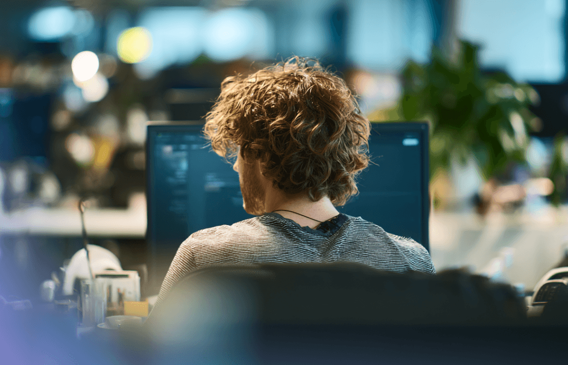 Blick über die Schulter auf einen Entwickler mit lockigem Haar, der in einem modernen Büro an einem Computer arbeitet.